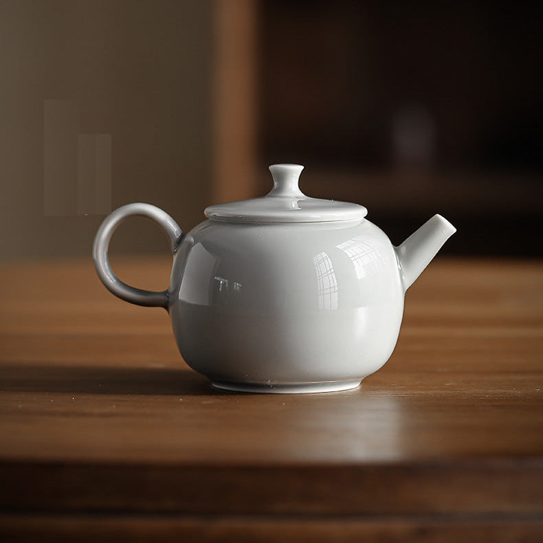 A minimalist white ceramic teapot with a rounded body, lid, and loop handle, resting on a wooden table with a blurred background.
