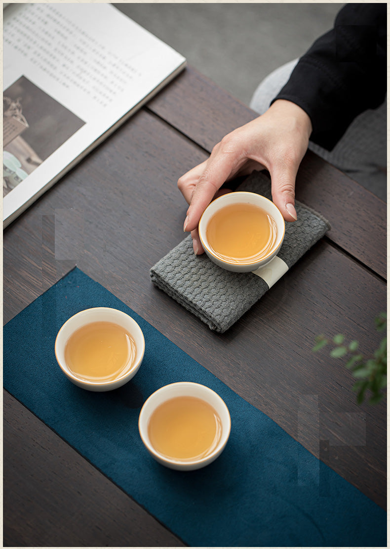 A hand holds a small white cup of amber tea on a gray textured cloth over a dark wooden table, with two more tea cups on a blue runner nearby and an open book in the background.
