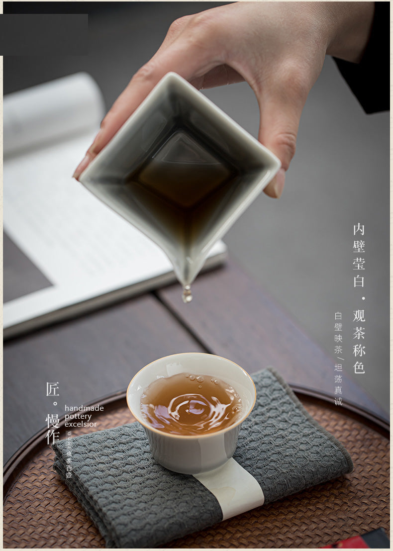 A hand holds a square white ceramic pouring vessel over a small white cup containing amber tea, set on a textured gray cloth over a woven tray. An open book lies blurred in the background, with Chinese text printed along the right side.