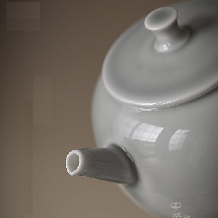 A close-up of a glossy white ceramic teapot with a lid and small spout, photographed against a warm taupe background. Chinese characters are printed vertically on the lower right side.