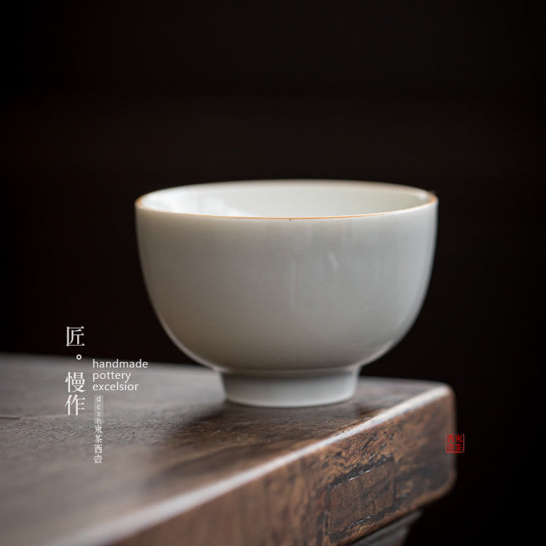 A handmade ceramic tea bowl with a pale celadon glaze sits on a dark wooden surface against a dark background. The image features Chinese calligraphy text on the left and a brand logo in the upper right corner.