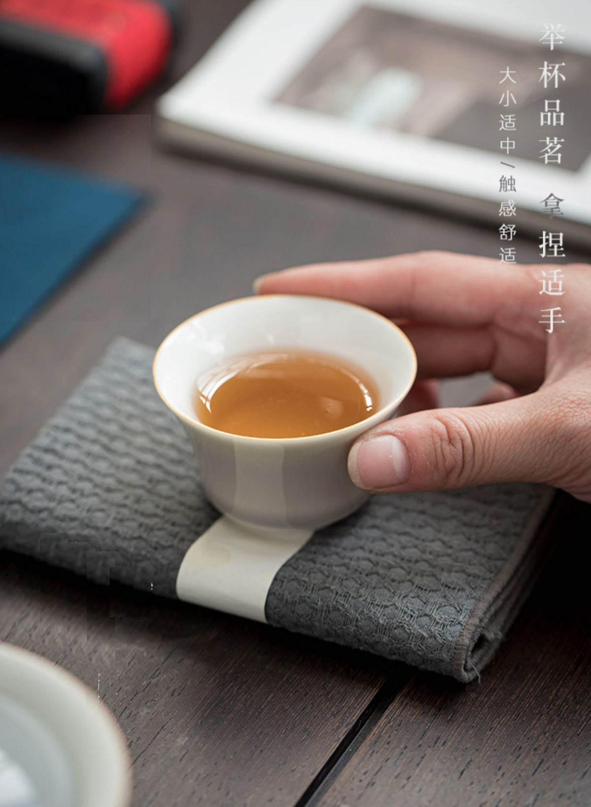 A hand holding a small white ceramic tea cup filled with amber tea, resting on a gray textured cloth over a dark wooden table. Chinese text appears vertically on the right side.