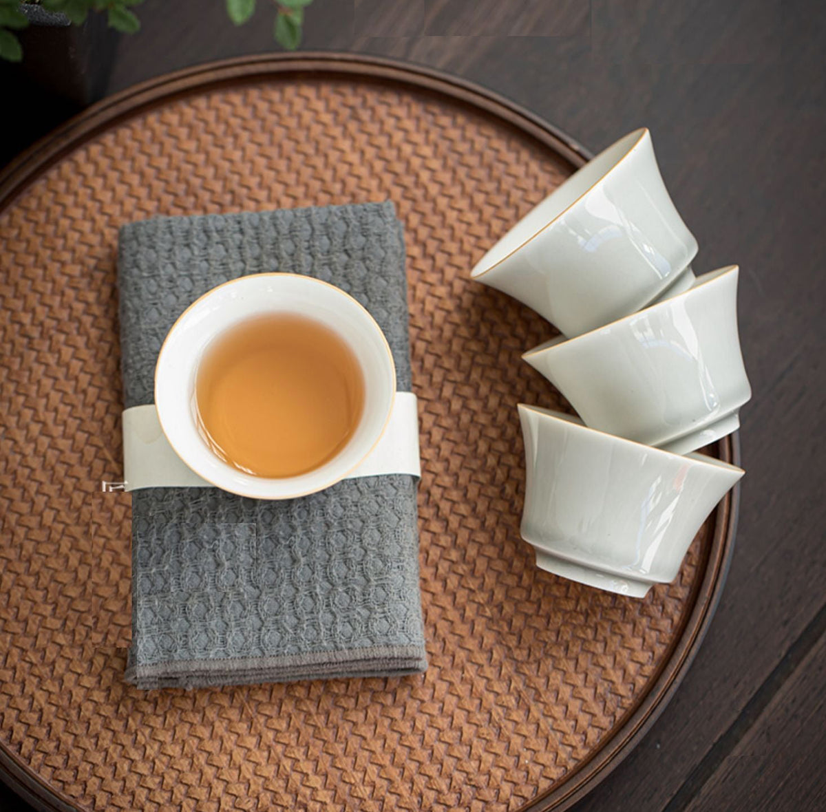 A tea set arrangement on a woven round tray, featuring a white ceramic teacup with amber tea and a folded gray cloth, alongside three stacked white ceramic cups, on a dark wooden surface.