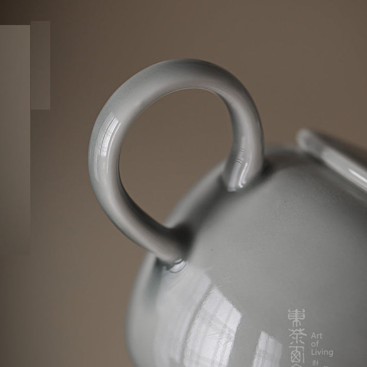 Close-up of a glossy light gray ceramic teapot with a loop handle, set against a warm brown background. Chinese characters are printed on the side.