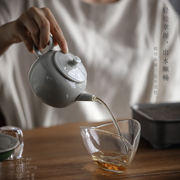 A person in a white shirt pours amber tea from a matte gray ceramic teapot into a clear glass公道 cup on a wooden table. The scene has a warm, soft-focus aesthetic with Chinese text overlays on the right and a brand logo on the left.
